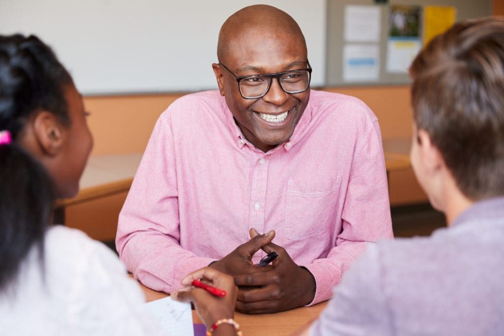 male-high-school-tutor-sitting-with-students-at-WM5F4CS-1024x683